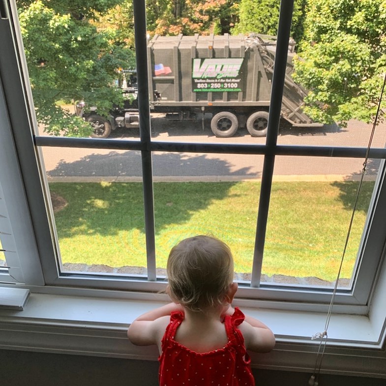 A young child in a red outfit looks out a window at a Value Waste garbage truck parked outside.