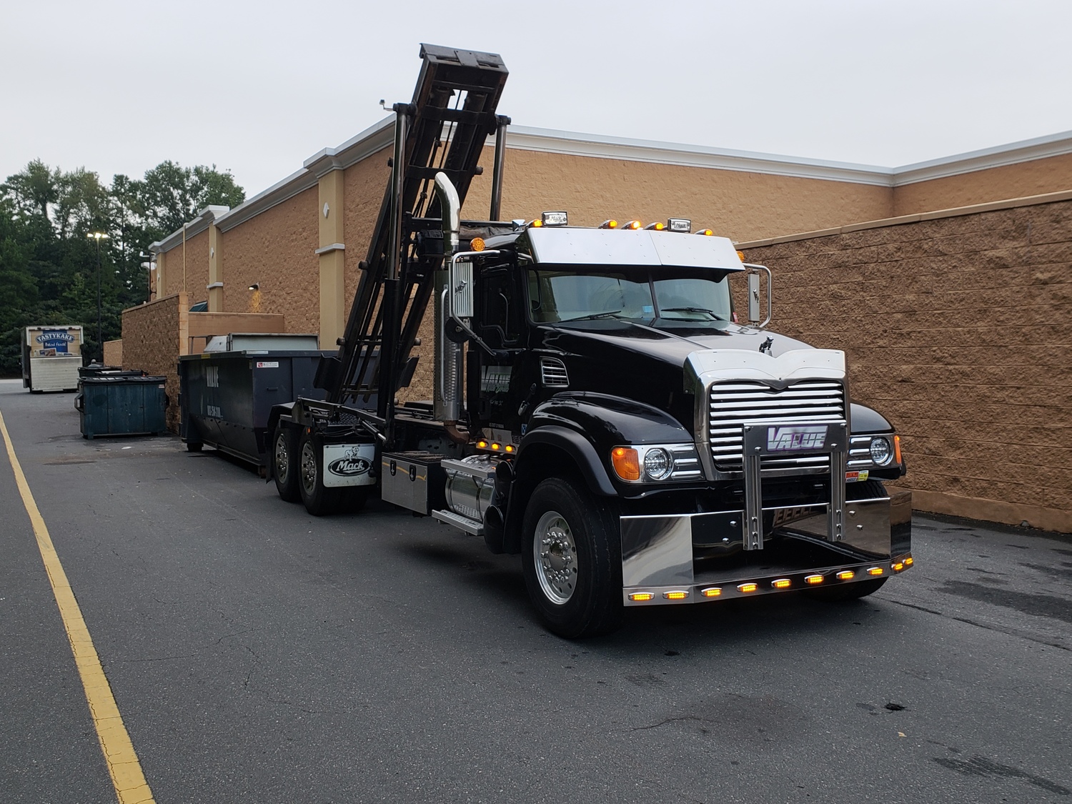 A black roll-off truck with the Value Waste logo, parked in front of a store.