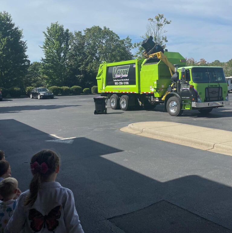 A green garbage truck with the Value Waste logo, picking up a trash can while a group of children watch.