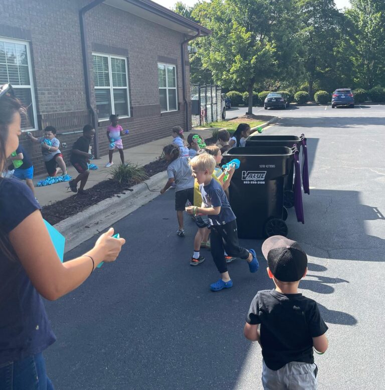 Children playing outdoors with water guns on a sunny day near a building and a black trash bin.