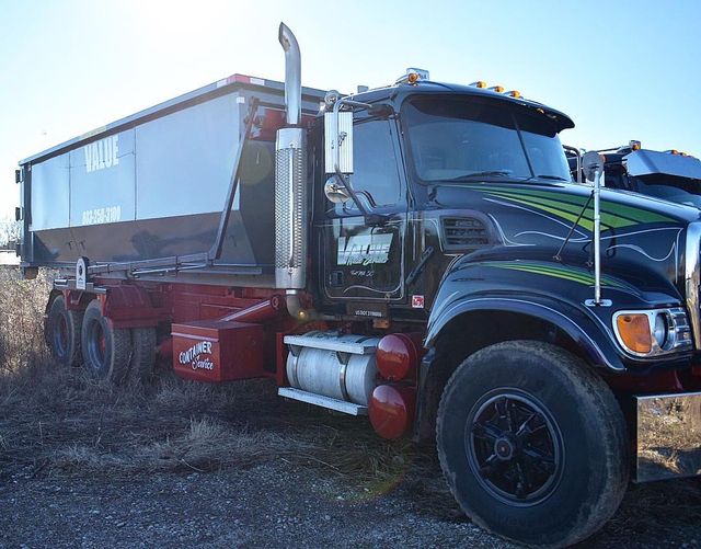 A black roll-off truck with the Value Waste logo, parked on a dirt road.