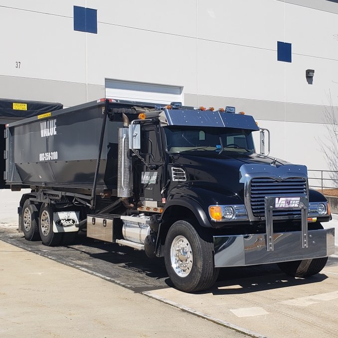 A black roll-off truck with the Value Waste logo, parked in front of a warehouse.