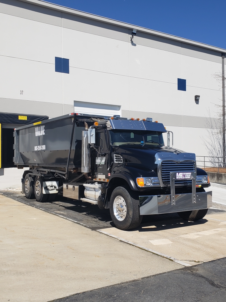 A black roll-off truck with the Value Waste logo, parked in front of a warehouse.