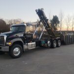 A black roll-off truck with the Value Waste logo, lifting a dumpster filled with debris.