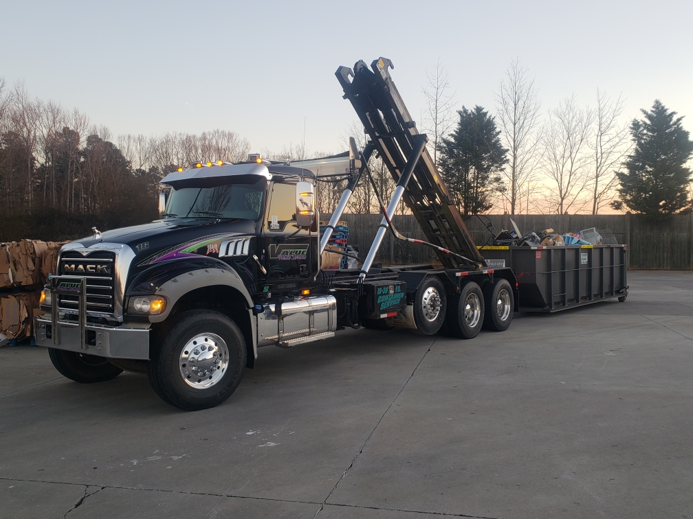 A black roll-off truck with the Value Waste logo, lifting a dumpster filled with debris.