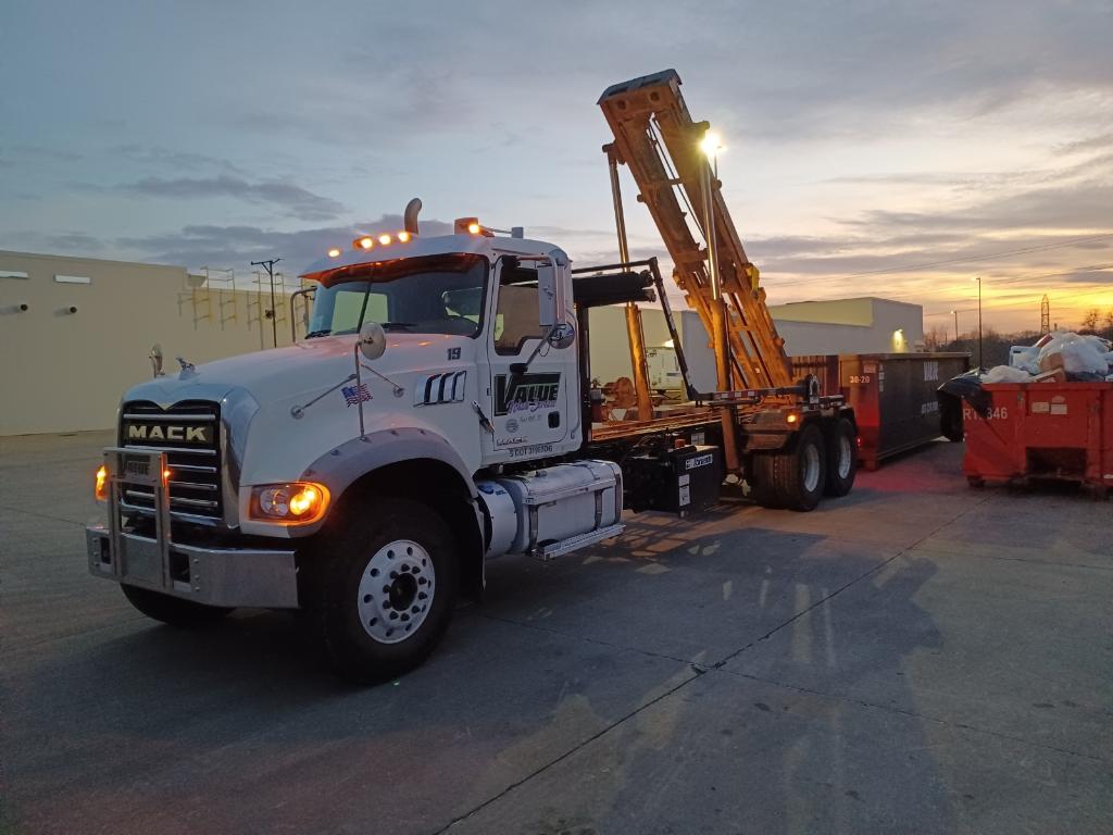 A white roll-off truck with the Value Waste logo, lifting a dumpster at sunset.