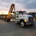 A white roll-off truck with a dumpster in the background, parked at sunset.