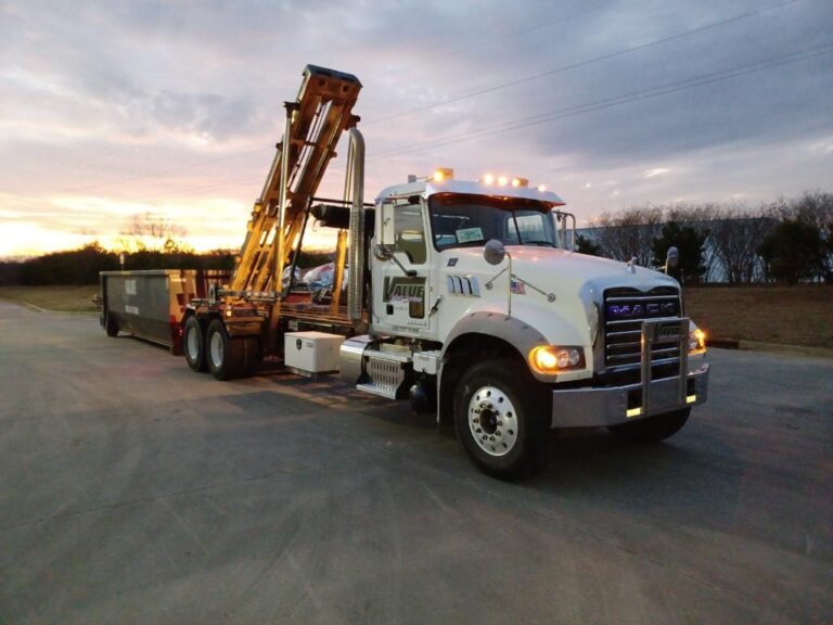 A white roll-off truck with a dumpster in the background, parked at sunset.
